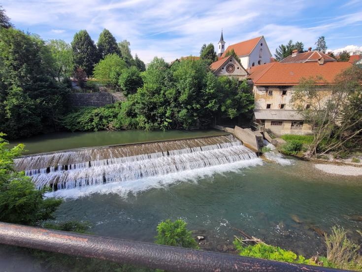 The scenic beauty of Škofja Loka, Slovenia, where the FBI4EU project partners gathered for their initial meeting, set the perfect backdrop for collaborative discussions and networking. A picturesque view of a small waterfall and river in Škofja Loka, Slovenia, surrounded by lush greenery and historic buildings with red-tiled roofs. The clear blue sky and serene landscape provide a peaceful atmosphere.