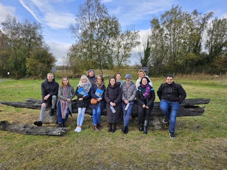 ADAPTO Project Partners Visit Cabragh Wetlands A group of ADAPTO project partners sits together on a large wooden bench outdoors at Cabragh Wetlands, Ireland. Behind them are green trees and an open field. The group, dressed warmly for autumn, is smiling and holding informational brochures.