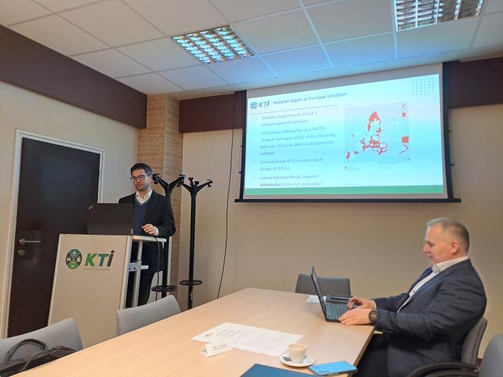 A man is presenting a powerpoint and another one is taking note on his computer