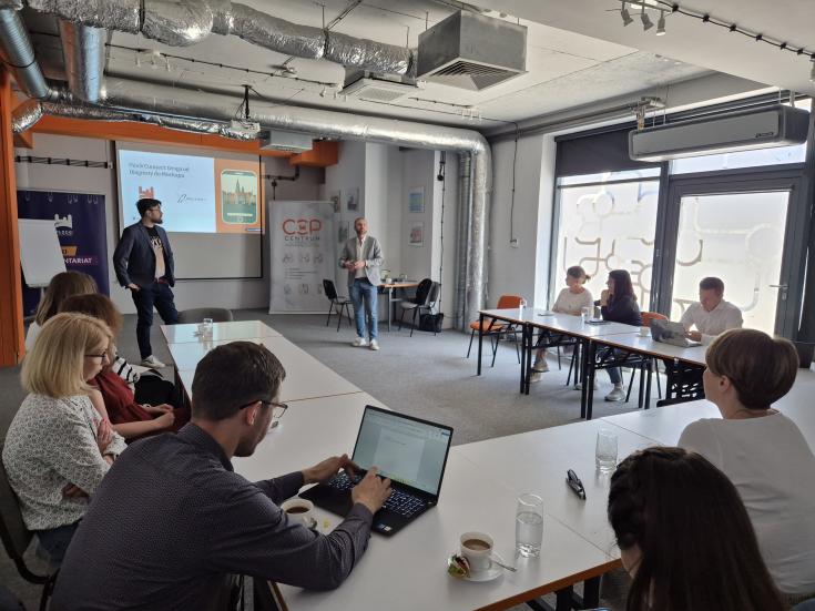 Płock Stakeholders Meeting – Presentation of “Płock Connect” Migrant App People attend a meeting where two men present the “Płock Connect” migrant app. Participants sit around tables with laptops and notes.