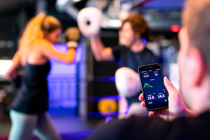 In front a man holding a phone showing data. In the backround two women are boxing.