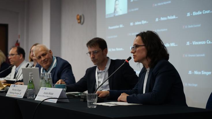 A woman speaks into a microphone while seated at a table with other speakers during a professional conference. One of the male speakers is gazing in her direction. Behind her is a screen projection of others connected to the conference via Zoom.