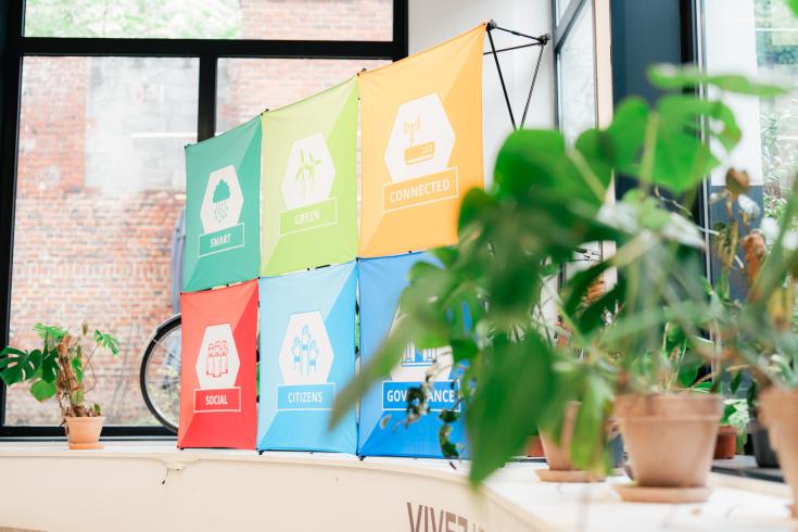 Projects banner with green plant Six colorful Interreg Europe project logos are positioned in a printed banner, sitting in front of a window in an industrial corporate space. A plant sits in the foreground of the image, closest to the camera.