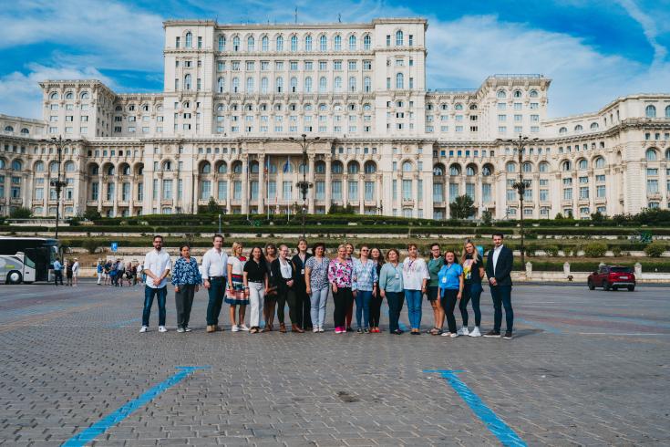 A group of participants from the SHARING project event poses in front of the imposing Palace of the Parliament in Bucharest, Romania, under a bright blue sky. The group consists of men and women standing on a cobblestone square, facing the camera, with some smiling and engaging with each other. In the background, the massive white neoclassical facade of the building stretches across the scene, adding grandeur and context to the gathering.