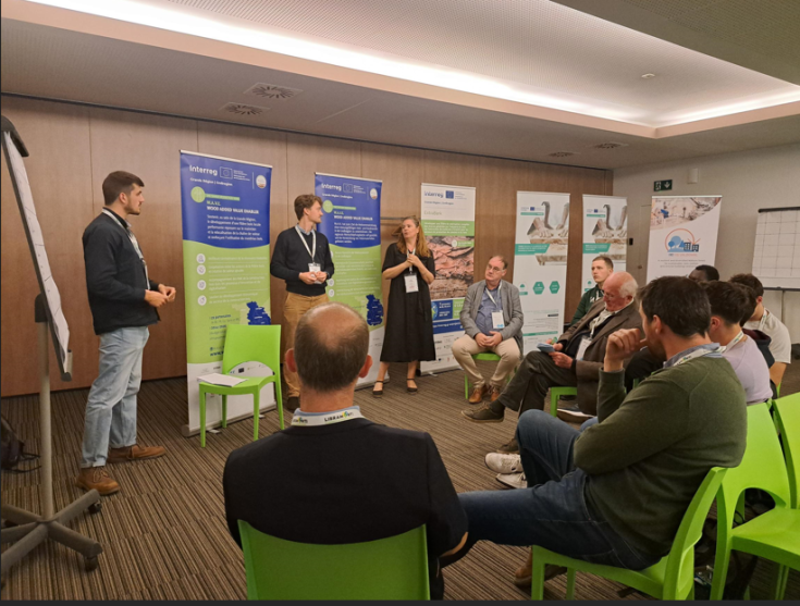 Sharing Experiences at Les Rencontres Filière Bois Participants engaged in a discussion at Les Rencontres Filière Bois event, with attention on a woman speaker standing near banners showing Interreg project logos.