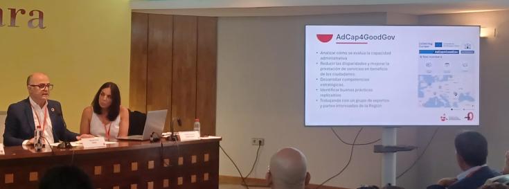 FMRM & AdCap4GoodGov at the conference ‘ICT Priorities in the Murcia Public Administrations in view of the Next Generation Funds’. The image shows a conference setting, where two individuals are seated at a table T A screen on the right displays a slide titled AdCap4GoodGov,