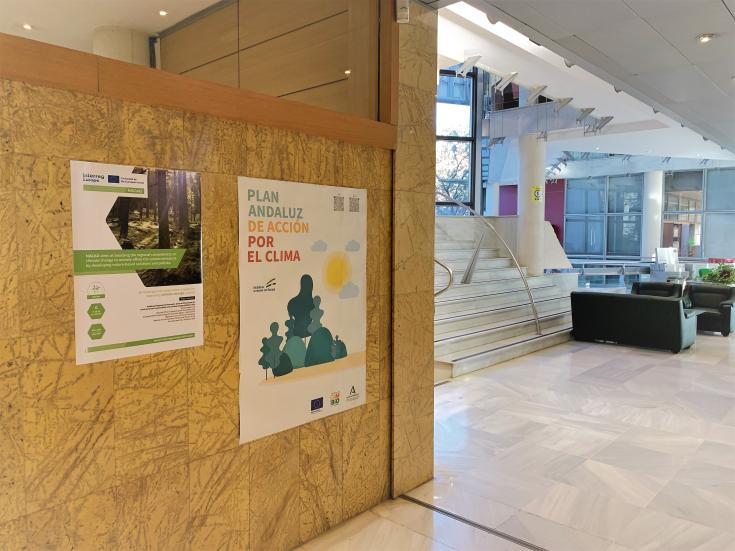 Poster on a wooden wall in main entrance of the building, behind corner are white stairs.