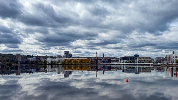 A dramatic cloudy sky over a modern cityscape reflected in calm water. Buildings, including colorful structures and a church spire, line the waterfront. A red buoy floats in the foreground