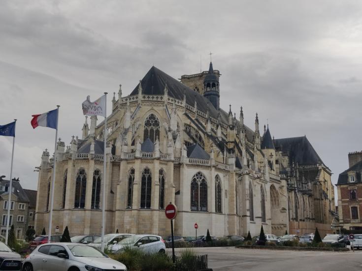 A Gothic-style cathedral with pointed arches, ornate spires, and large stained glass windows, seen from a plaza with parked cars. Three flags—EU, French, and a local flag—stand in the foreground, suggesting a European town setting