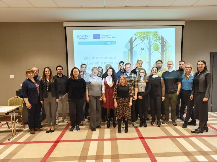 Participants of the 2nd International Partnership Meeting of the FBI4EU project, held in Rzeszów, gathered for a group photo to commemorate the event. A group of individuals standing in a conference room in front of a projector screen displaying a slide for the "2nd International Partnership Meeting" of the FBI4EU project. The background slide shows the Interreg Europe logo and an illustration of a forest.