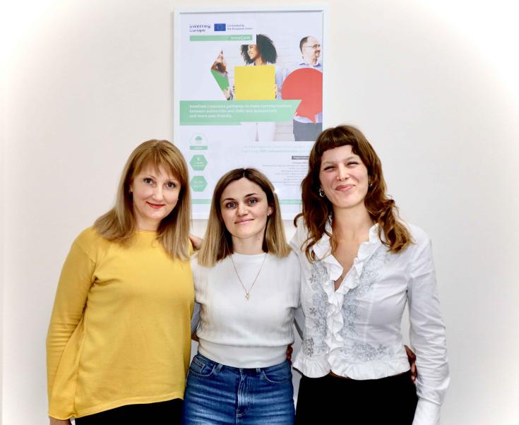 Three women pose in front of the project poster