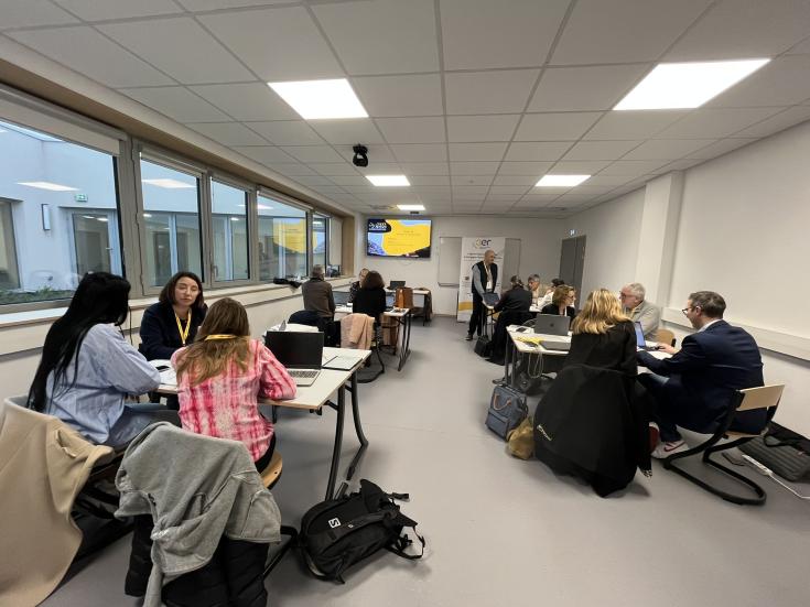 A group of people are engaged in discussions around tables in a bright, modern classroom. Some are working on laptops while a presenter addresses the group at the front.