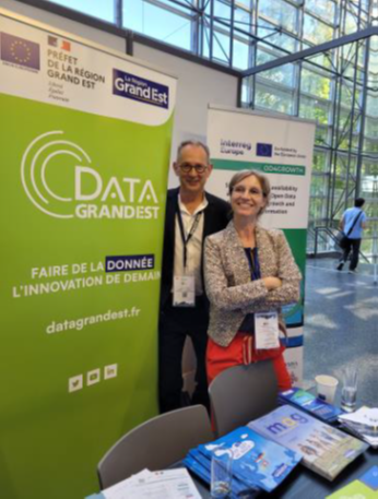 Event photo It's a photo of the stand shared between our partner and other stakeholders. In the center, it's possible to see a man and a woman posing for the photo. In the right side of the photo, we see a green banner with relevant information about the stand and the event.