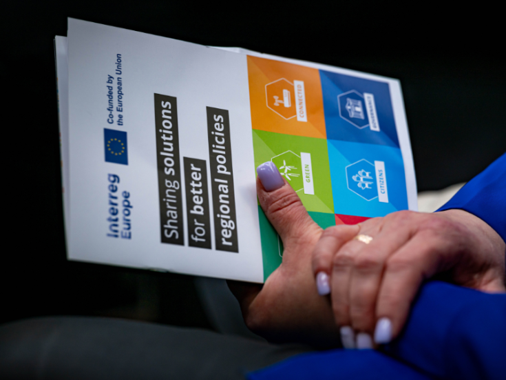 Interreg Europe branding A person with purple nails holds a brochure with the text "Sharing solutions for better regional policies"
