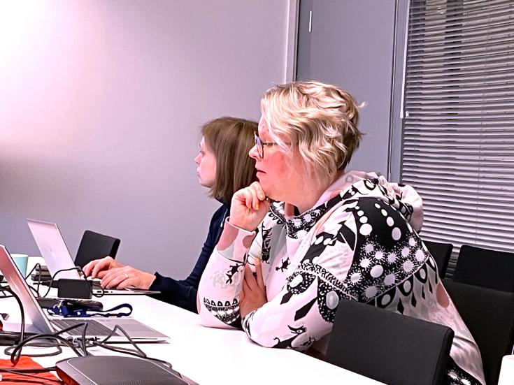 Light-haired woman sitting beside the meeting room table looking down to her laptop and giving presentation.