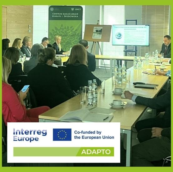 ADAPTO Interreg Europe Meeting in a Modern Conference Room in Poland A group of professionals seated around a large conference table in a well-lit meeting room, attentively listening to a presenter at the front. The room features modern furnishings and several attendees are looking at digital screens.