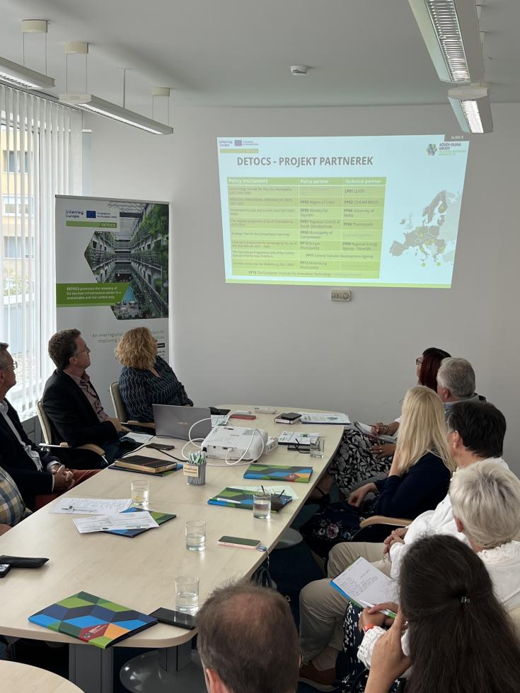 Numerous people sat around a large rectangular table watching a project presentation on a display projector.