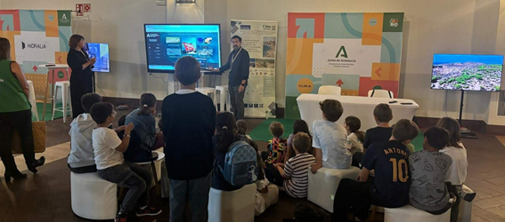 A man lectures in front of a large screen to a group of schoolchildren sitting on chairs and stools.