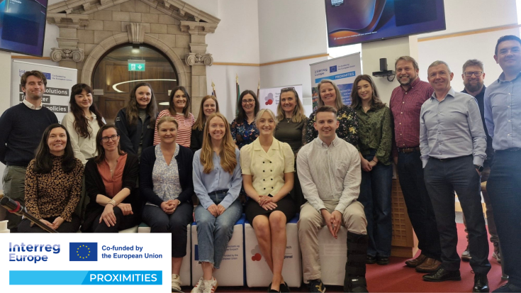 PROXIMITIES Stakeholders Gather in Waterford city! The image shows a group of around 20 people posing for a photo at a professional event, likely related to the Interreg Europe PROXIMITIES project, as indicated by the banners in the background. The group is a mix of men and women in business casual attire, standing and sitting in two rows in a formal setting with stone architecture, flags, and presentation screens, creating a collaborative and official atmosphere.