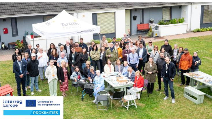 Group photo of PROXIMITIES study visit participants and local residents gathered outdoors at Arus Mhuire, an age-friendly housing scheme in Cork. People are smiling and standing or sitting around picnic tables, under a Tuath Housing canopy. PROXIMITIES and Interreg Europe logos are visible in the foreground.