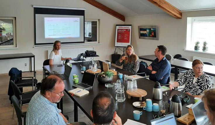 A group of stakeholders engaged in a discussion during the GIFT project Regional Stakeholder Group meeting in Fryslân, July 2024. A presentation is displayed on a screen, and participants are seated around a conference table with laptops, coffee, and refr