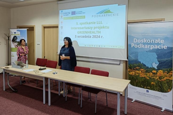 A woman standing behind a table speaks in a closed room. Behind a powerpoint screen and on the side another woman standing and listening.