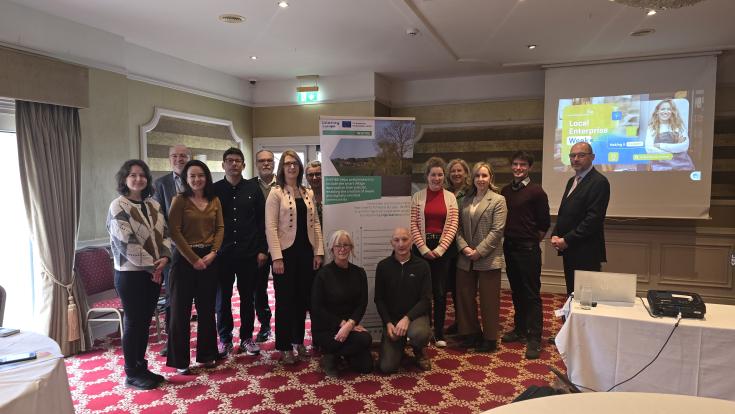 In the picture, a diverse group of men and women from the INSPIRE project stands together, posing for a photo. They are gathered after a Regional Stakeholder meeting at a facility in Spruce & Willow, located in Ardpatrick, Co. Limerick. The group smiles and stands proudly in front of a large banner displaying the INSPIRE project logo, highlighting their collaboration and the purpose of the event. The setting appears professional, with the modern facility in the background, offering a sense of teamwork.