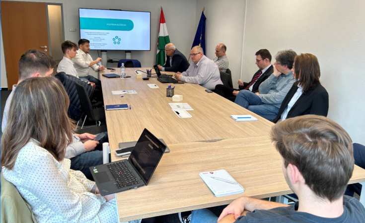 The photo shows a group of people sitting around a table, engaged in discussion. It appears to be a meeting or workshop related to the INSPIRE Interreg Europe Project.  In the background, In the background, the European Union flag and the Hungarian flag are visible. There’s also a projector displaying slides, suggesting that a presentation is taking place.