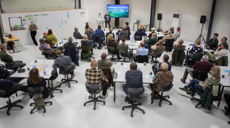Forestry Conference on Sustainable Management in Norddjurs A forestry conference in Norddjurs, Denmark, with participants seated at round tables listening to a speaker presenting on a large screen. A facilitator is writing on a whiteboard in the background. The event takes place in a modern indoor setting with professional equipment.