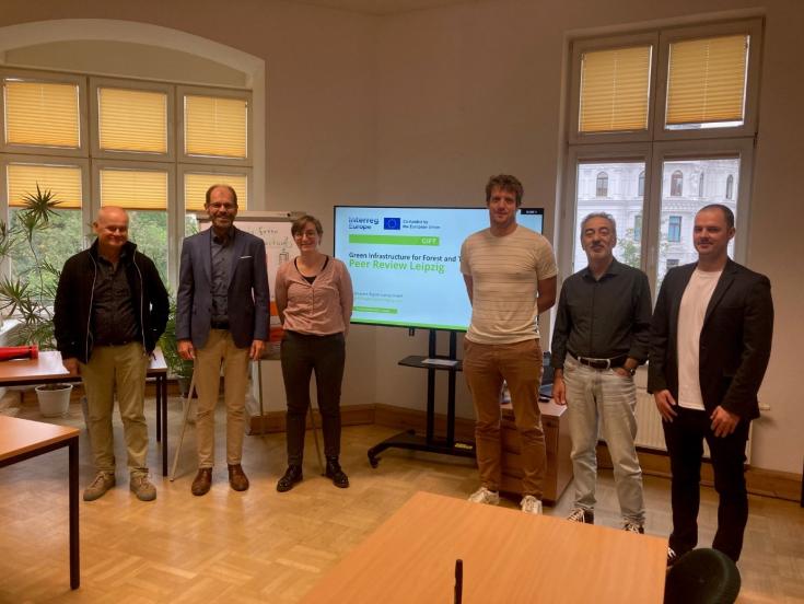 Group of six participants at the GIFT Project peer review session in Leipzig, standing in front of a presentation screen displaying “Green Infrastructure for Forest and Territory - Peer Review Leipzig.” Participants include representatives from SMEKUL, Dübener Heide Nature Park, and other project partners.