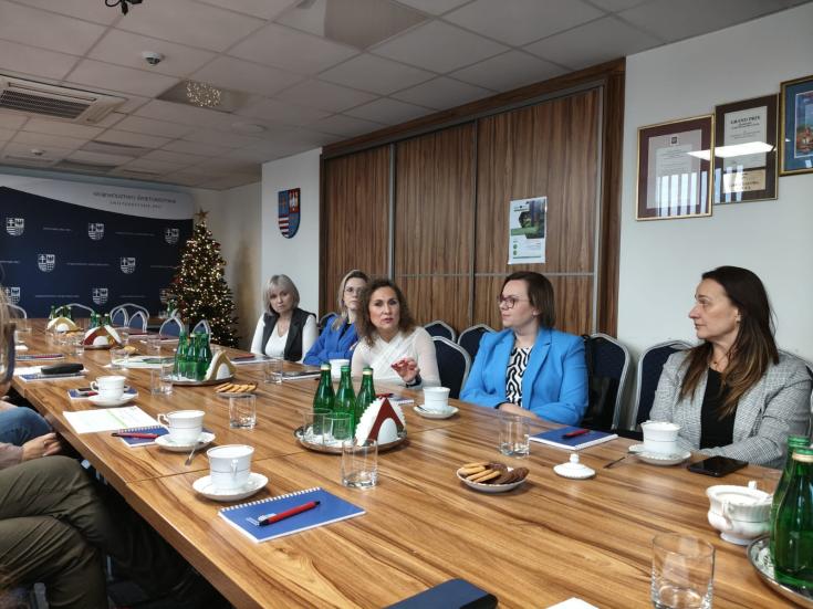 People in a brown coloured meeting room around long table. One women with white jacket raises a question.