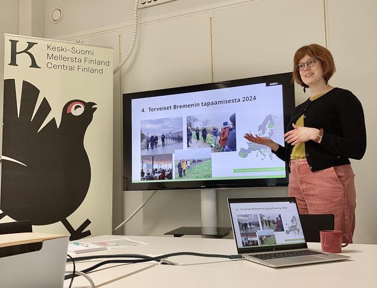 Woman giving a presentation in front of the screen. She has red mid-lenght hair, green t-shirt and marsala pink trousers.