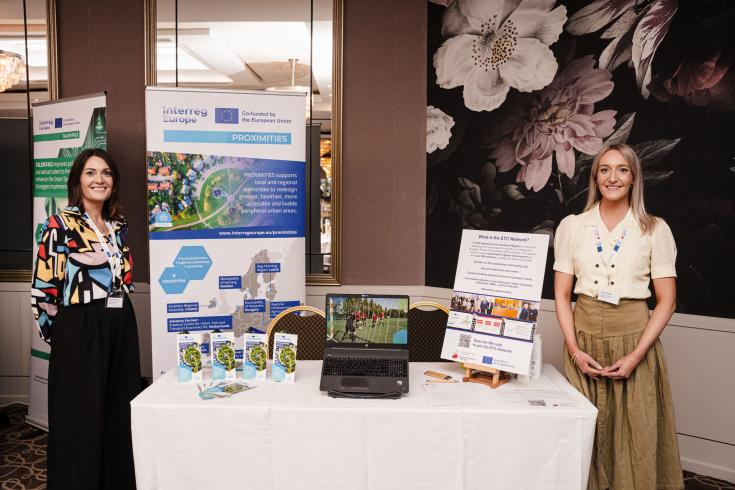 PROXIMITIES Highlighted in Europe Day Celebrations Two women stand on either side of a promotional display table for the Interreg Europe PROXIMITIES project at an indoor event. Behind them are banners showcasing the PROXIMITIES project and the ETC Network. On the table are brochures, a laptop playing a video, and informational materials. The setting appears to be a conference or exhibition space with floral wallpaper and elegant decor.