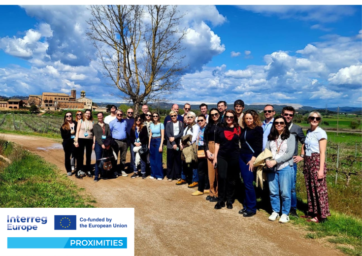 PROXIMITIES partners during the study visit to Manresa, March 2025 A group of PROXIMITIES project partners from across Europe pose together outdoors during a study visit to Manresa, Spain. They stand on a rural path with green fields, a large tree, and a historic building in the background under a partly cloudy blue sky. Interreg Europe and PROXIMITIES logos appear in the bottom-left corner.