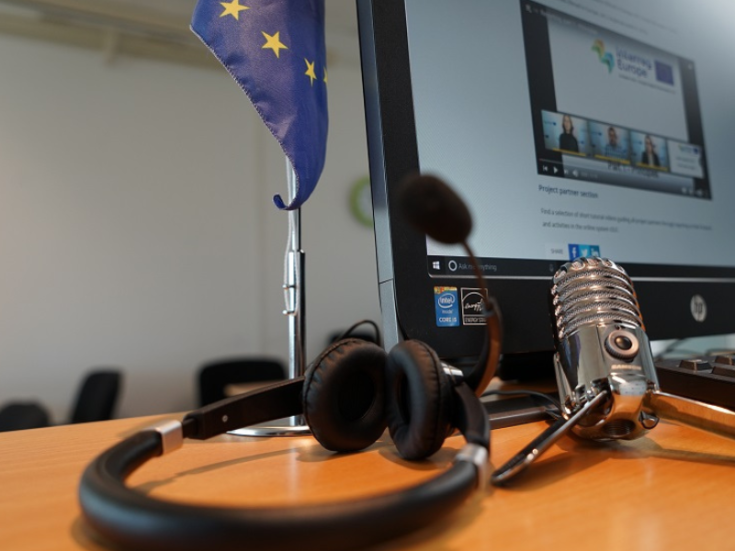 Professional tools for creating a video Close-up of a desk setup for virtual meetings or online collaboration. The desk features a European Union flag, a computer monitor displaying a video call or webinar interface, a pair of black headphones, and a vintage-style microphone.