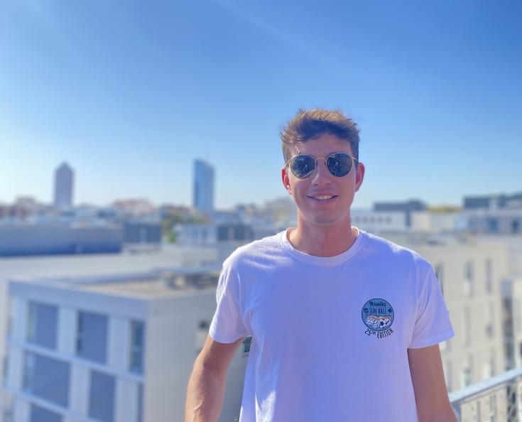 Dark-haired student boy with sun glasses and white T-shirt smiles at the rooftops of Lyon.