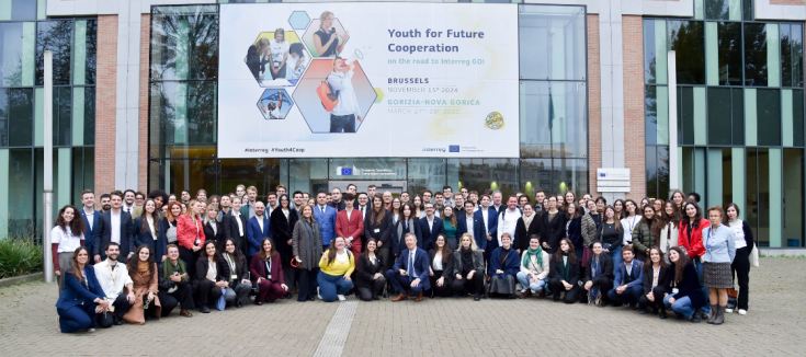 A large group of young people poses for a group photo in front of a modern glass-fronted building. Above them, a large banner reads "Youth for Future Cooperation – on the road to Interreg GO!", with event details indicating gatherings in Brussels (November 15, 2024) and Gorizia-Nova Gorica (March 27-28, 2025). The banner also features images of young professionals engaging in discussions and collaborative activities. The setting appears to be an official EU institution, with the European Commission logo vis