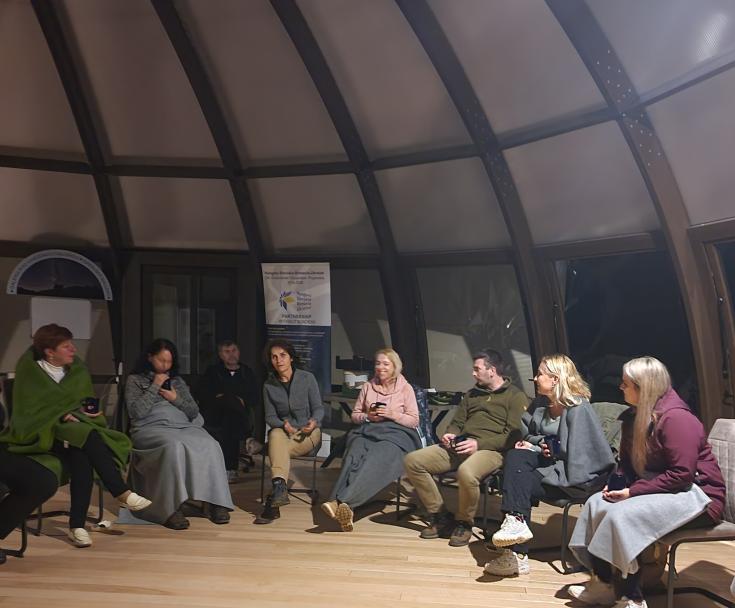 A group of persons in a room sitting in circle, nighttime.