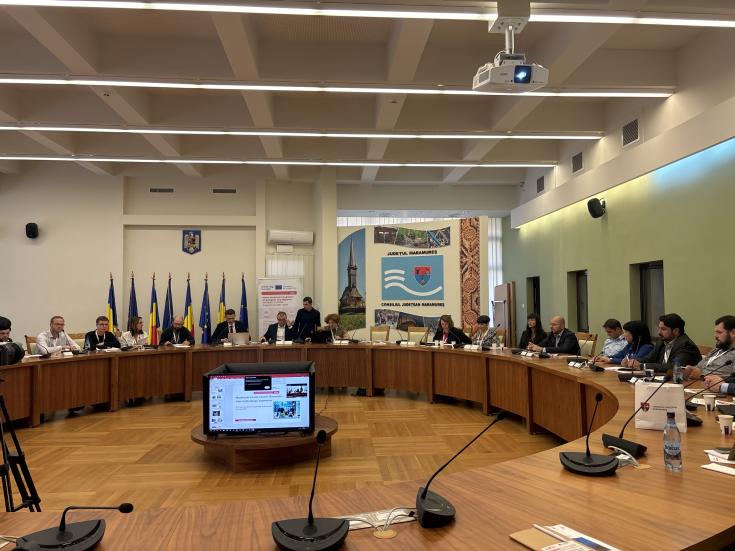 4th Transnational Learning Meeting (Romania) People sitting in a large room at desks arranged in a circle, with a presentation projected on a display in the centre.