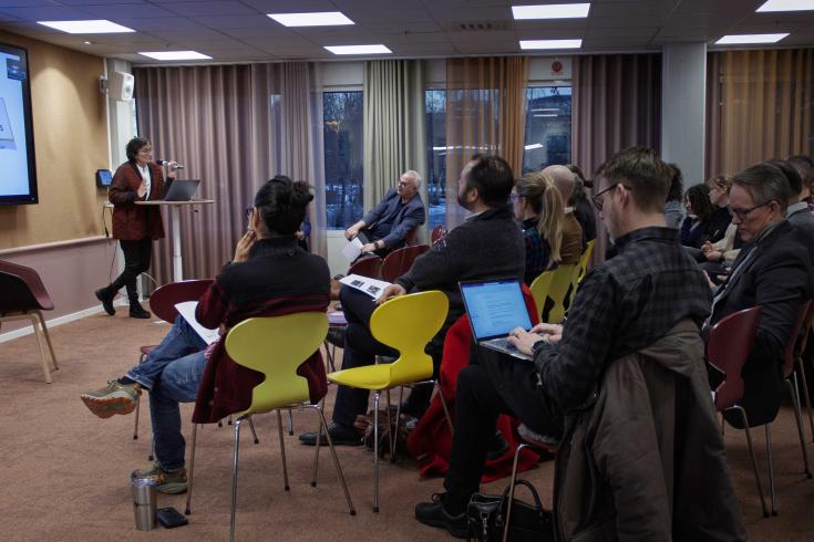 A presenter speaks at a podium to an audience seated in colourful chairs, some taking notes or using laptops.