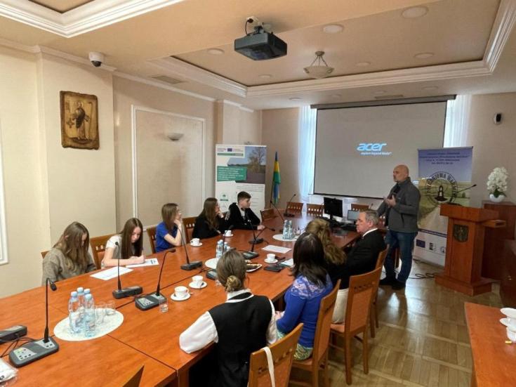 In the picture, the youngest members of the municipality’s Youth Council in Olsztyn, Poland, are positioned at the center, actively engaged around a rectangular table in an institutional setting. They are focused on taking notes and participating in the discussion. On one side of the group, Mr. Tomasz Piłat, the partner leader of the INSPIRE Project and a representative from the LAG Association "South Warmia," is interacting with them.