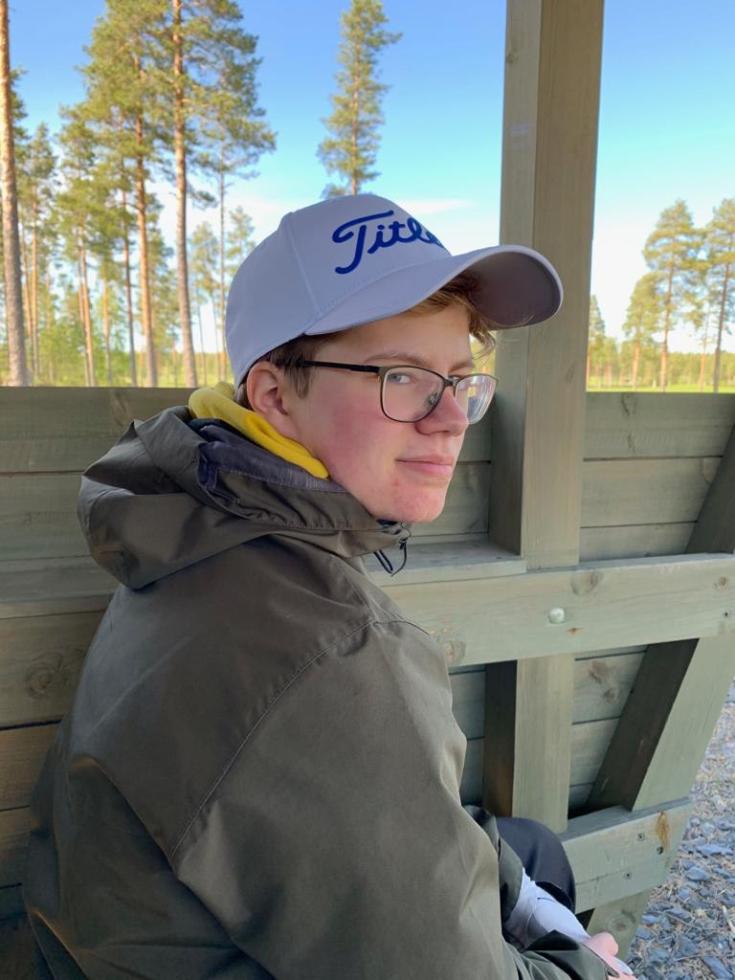 A boy with white cap, green jacket and glasses sitting on a bench beside the forest.