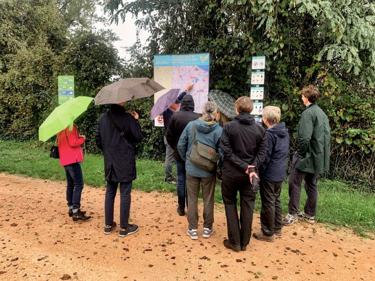 A group of people holding umbrellas stand outdoors in front of an information board and signage, examining a map and illustrations on biodiversity. The group is dressed in rain jackets, indicating rainy weather, with green foliage and a gravel path in the background. The scene appears to be part of an outdoor learning session or study visit focused on environmental conservation and green infrastructure.