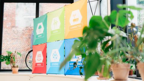 Six colorful Interreg Europe project logos are positioned in a printed banner, sitting in front of a window in an industrial corporate space. A plant sits in the foreground of the image, closest to the camera.
