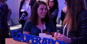 Two people talk and laugh during an event standing next to a table with the word cooperate on it