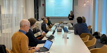 The image shows a professional meeting taking place in a modern conference room. A group of people is seated around a long wooden table, working on laptops, taking notes, or listening to a presentation. The presenter, a woman dressed in black and white, is standing at the front of the room, speaking while gesturing towards a projector screen.