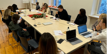 Meeting participants seated around tables arranged together. All looking towards the screen on which the presentation is projected. In the corner of the room to the right of the screen, a whiteboard is visible.