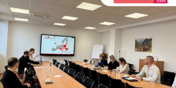 Meeting participants sitting at tables and looking at a screen visible at the end of the room. A presentation is projected on the screen.