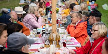 A large group of people enjoying lunch at a long table in a park.