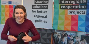 person smiling and standing in front of a colourful Interreg Europe stand and holding headphones